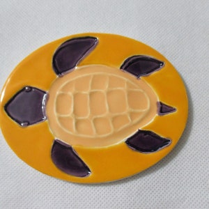 May include: A ceramic turtle coaster with a yellow background and purple accents. The turtle's shell is a light orange color.
