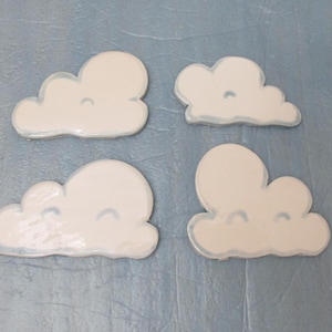 Può includere: Quattro ornamenti in ceramica bianca a forma di nuvola con contorni blu e occhi dipinti. Le nuvole sono disposte a scalare su uno sfondo blu chiaro.