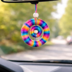 May include: A vibrant, handmade car air freshener with a rainbow yarn design. The circular charm, with a smaller inner circle, hangs from a rearview mirror. The yarn is a mix of pink, yellow, blue, and purple, creating a bright, eye-catching accessory for the car.