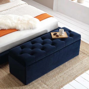 May include: A navy blue velvet storage ottoman with tufted buttons. The ottoman is sitting on a woven rug in front of a bed with white bedding and an orange throw blanket.