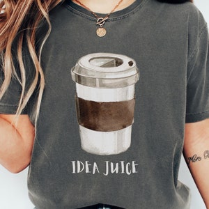 Könnte beinhalten: Dunkelgraues T-Shirt mit einer Aquarellillustration einer Kaffeetasse mit brauner Manschette. Der Text "IDEA JUICE" ist darunter in Weiß gedruckt. Das Shirt hat einen bequemen, lässigen Stil.