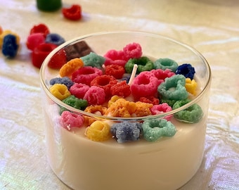 May include: A white candle in a clear glass container, decorated with colorful, cereal-shaped wax pieces. The wax pieces are in various colors, including pink, blue, green, yellow, and orange. A white wick is centered in the candle.