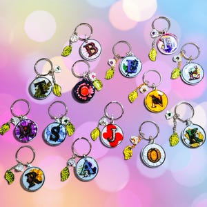 May include: A collection of circular keychains featuring individual letters of the alphabet. Each keychain includes a metal ring, a small green leaf charm, a white flower charm, and a resin disc containing a letter with colorful floral accents.