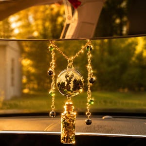 Può includere: Un ornamento da appendere allo specchietto retrovisore dell'auto con una catenella dorata e perline verdi e nere. Un ciondolo circolare in vetro mostra la sagoma nera di un cervo in una foresta, collegato a una piccola fiala di vetro con scaglie dorate.