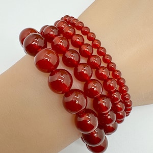 May include: Three red beaded bracelets of varying sizes are stacked on a wrist. The beads are round and have a glossy finish. The bracelets are a deep, rich red color, and the light reflects off the beads.