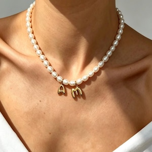May include: A necklace featuring a strand of white, round pearls with two gold-toned metal charms hanging from the centre. The charms are shaped like the capital letters A and M, each suspended from a small gold ring attached to the pearl strand.