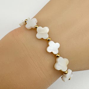 May include: A white clover-shaped bracelet with gold-colored beads. The bracelet is made of white mother-of-pearl and gold-colored metal. The bracelet is shown on a wrist.