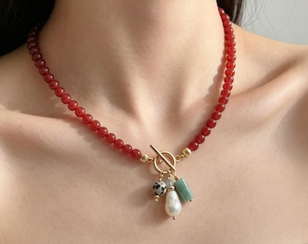 May include: A necklace featuring a strand of small, round, deep red beads with a gold-toned toggle clasp. Three charms hang from the center: a small round bead with black spots, a white baroque pearl, and a rectangular green stone bead.
