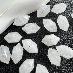 May include: A collection of clear, faceted quartz crystal beads. Each bead is a hexagonal shape with pointed ends. Some beads have a hole drilled through them. The beads are scattered on a black textured surface.