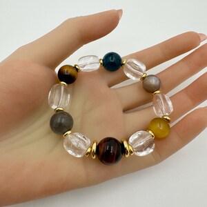 Pode incluir: Uma pulseira de contas com uma variedade de pedras preciosas em tons claros, castanhos, amarelos e azul escuro. A pulseira inclui detalhes metálicos dourados. A pulseira é segurada em uma mão.