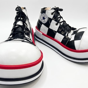 Puede incluir: Un par de zapatillas altas con un patrón de cuadros en blanco y negro, cordones negros y una puntera blanca redondeada de gran tamaño. Los zapatos tienen una suela de plataforma gruesa con una franja roja y el logo Converse All Star.