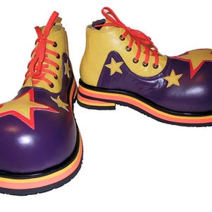 Puede incluir: Un par de zapatos de payaso con base morada, aplicaciones de estrellas amarillas y ribetes rojos. La parte superior es amarilla con cordones rojos y ojales metálicos. La suela consta de capas apiladas en negro, naranja y amarillo con una puntera redondeada.