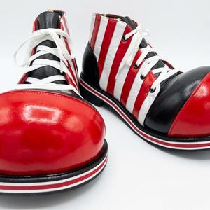 Puede incluir: Un par de zapatos de payaso de gran tamaño con una puntera redondeada de color rojo brillante, una parte superior a rayas verticales blancas y negras y cordones blancos. Tienen una suela negra gruesa con una fina raya roja y blanca.