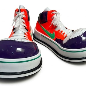 Puede incluir: Un par de zapatillas altas con una puntera morada redondeada de gran tamaño, una parte superior de color naranja brillante y un logotipo swoosh verde. Los zapatos tienen cordones blancos, una puntera perforada blanca y una suela de plataforma blanca gruesa con un borde negro.
