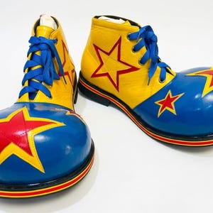 Puede incluir: Un par de zapatos de payaso de gran tamaño con una parte superior amarilla y una puntera azul redondeada. Los zapatos están decorados con estrellas rojas con bordes amarillos, cordones azules y una suela negra con rayas rojas y amarillas.
