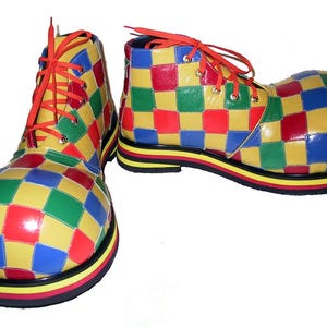 Puede incluir: Un par de zapatos de payaso de gran tamaño con un vibrante patrón de cuadros en rojo, amarillo, azul y verde. Los zapatos tienen cordones de color naranja brillante, ojales metálicos y una suela gruesa con rayas horizontales negras, amarillas y rojas.