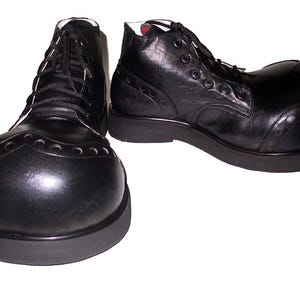 Puede incluir: Un par de zapatos de payaso de cuero negro con una puntera redondeada de gran tamaño, cierre frontal con cordones negros y una suela de goma negra gruesa y plana. El diseño incluye detalles decorativos perforados en la puntera.