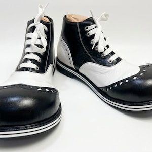 Puede incluir: Un par de botas con cordones en blanco y negro con punteras redondeadas y de gran tamaño. Las botas presentan un diseño bicolor, ojales metálicos plateados, cordones blancos y una suela superpuesta con rayas horizontales blancas y negras.