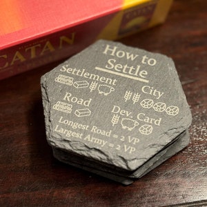 May include: A set of six black slate coasters with engraved instructions on how to settle a city in the board game Catan. The coasters feature icons for settlement, city, road, development card, longest road, and largest army.