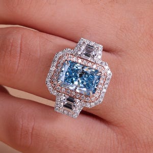 Pode incluir: Um anel de noivado deslumbrante com uma grande pedra preciosa retangular azul claro. O anel tem um design de halo duplo com um halo interno em ouro rosa e um halo externo em ouro branco, incrustado com pequenos diamantes. Diamantes de corte baguette adornam as laterais.