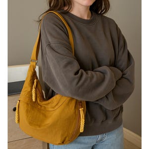 May include: Mustard yellow shoulder bag with a long adjustable strap. The bag has a zippered closure and decorative tassels. The person in the image is wearing a dark gray sweatshirt and blue jeans.