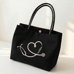 May include: Black tote bag with black handles and a white stethoscope design forming a heart shape. The word "custom" is written on the heart. The bag has a button closure.