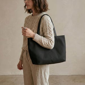 May include: A black tote bag with black leather handles. The bag is rectangular and appears to be made of a sturdy material. The bag is being carried over the shoulder.