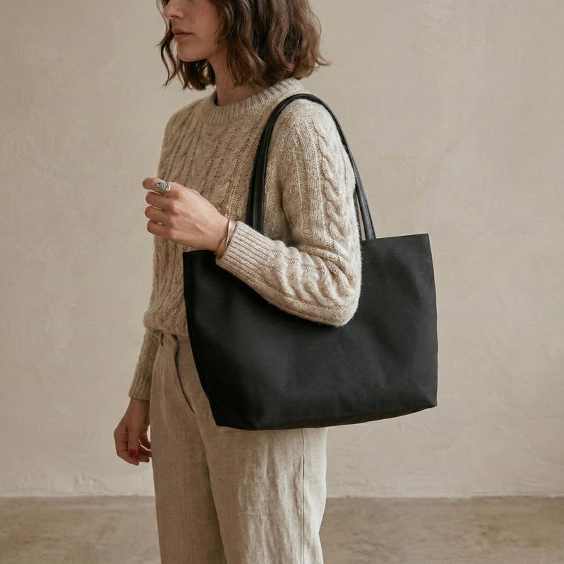 May include: A black tote bag with black leather handles. The bag is rectangular and appears to be made of a sturdy material. The bag is being carried over the shoulder.