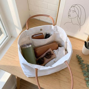 May include: A white canvas tote bag with brown leather handles, filled with various items. Inside are a green tumbler, a brown leather pencil case, a wallet, sunglasses, and other personal items. The bag sits on a wooden surface.