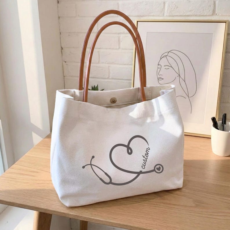 May include: White canvas tote bag with brown leather handles. The bag features a stethoscope design in the shape of a heart, with the word "custom" printed on the side. The bag is sitting on a wooden table.
