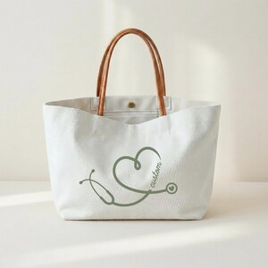May include: White canvas tote bag with brown leather handles. The bag features a green stethoscope design in the shape of a heart with the word "custom" in script. The bag has a button closure.
