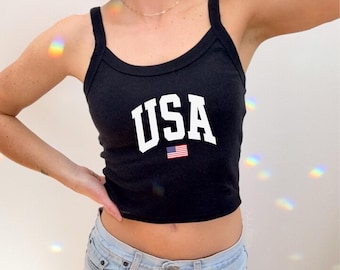 May include: Black tank top with the word "USA" in white block letters and a small American flag graphic. The top has a scoop neckline and spaghetti straps. The model is wearing light blue jeans.