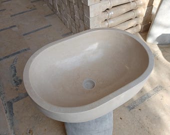 Puede incluir: Un lavabo ovalado de piedra beige con un orificio de desagüe central, apoyado sobre una base gris. La superficie lisa del lavabo contrasta con la textura más rugosa de la base. El fondo muestra balaustres de piedra apilados.