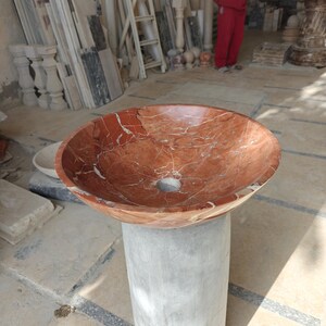 Puede incluir: Un lavabo de mármol rojo con vetas blancas, colocado sobre un pedestal de hormigón gris. La pila redonda tiene un orificio de desagüe central. El lavabo está en un taller con otros elementos arquitectónicos de piedra.
