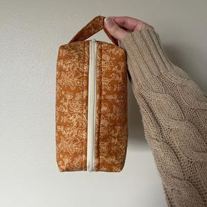 May include: A small, rectangular, brown zippered pouch with a white floral pattern. The pouch has a top handle and is made of quilted fabric.