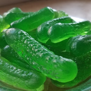 Op de afbeelding: Een bord met groene gummy snoepjes in de vorm van augurken. De snoepjes zijn doorschijnend en hebben een hobbelige textuur.