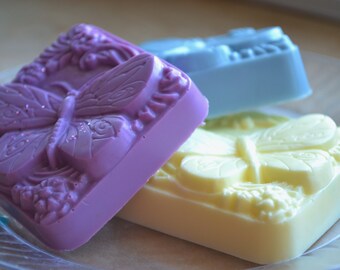 Könnte beinhalten: Drei handgemachte Seifenstücke mit Schmetterlingsmotiven. Die Seifenstücke sind lila, gelb und blau.