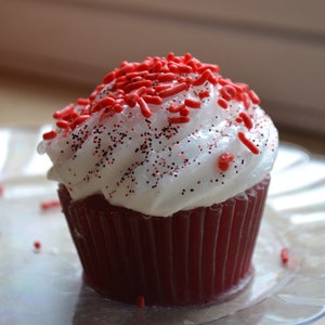 Puede incluir: Un cupcake de terciopelo rojo con glaseado blanco y chispas rojas. El cupcake está en un plato transparente.
