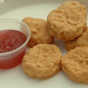 K&ouml;nnte beinhalten: Eine Gruppe von sechs goldbraunen, panierten H&auml;hnchen-Nuggets auf einem wei&szlig;en Pappteller mit beigem Muster, begleitet von einem kleinen, durchsichtigen Plastikbeh&auml;lter, der mit einer durchscheinenden roten So&szlig;e gef&uuml;llt ist.