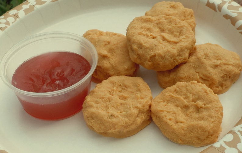 K&ouml;nnte beinhalten: Eine Gruppe von sechs goldbraunen, panierten H&auml;hnchen-Nuggets auf einem wei&szlig;en Pappteller mit beigem Muster, begleitet von einem kleinen, durchsichtigen Plastikbeh&auml;lter, der mit einer durchscheinenden roten So&szlig;e gef&uuml;llt ist.