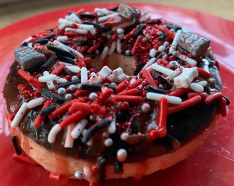 May include: A chocolate frosted donut with red and white sprinkles, red and black sprinkles, and silver glitter. The donut is on a red plate.