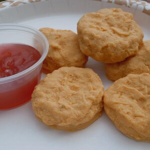 K&ouml;nnte beinhalten: F&uuml;nf goldbraune, runde, texturierte Lebensmittel, die H&auml;hnchen-Nuggets &auml;hneln, auf einem wei&szlig;en Teller neben einem kleinen, durchsichtigen Plastikbeh&auml;lter mit einer durchscheinenden, r&ouml;tlich-rosa Dip-Sauce.