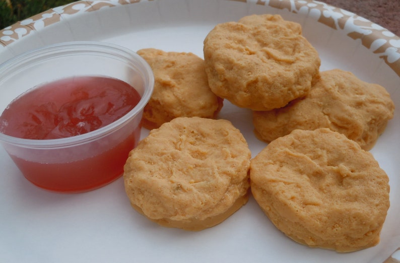 K&ouml;nnte beinhalten: F&uuml;nf goldbraune, runde, texturierte Lebensmittel, die H&auml;hnchen-Nuggets &auml;hneln, auf einem wei&szlig;en Teller neben einem kleinen, durchsichtigen Plastikbeh&auml;lter mit einer durchscheinenden, r&ouml;tlich-rosa Dip-Sauce.