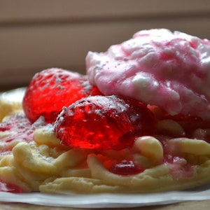 May include: A waffle dessert topped with pink whipped cream, red jelly candies, and powdered sugar.