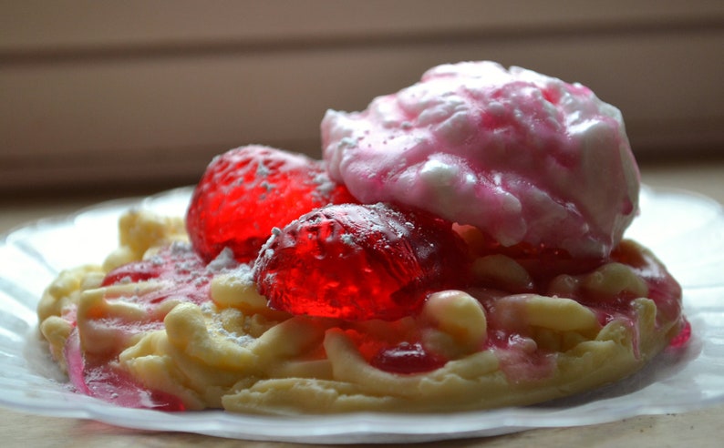 May include: A waffle dessert topped with pink whipped cream, red jelly candies, and powdered sugar.