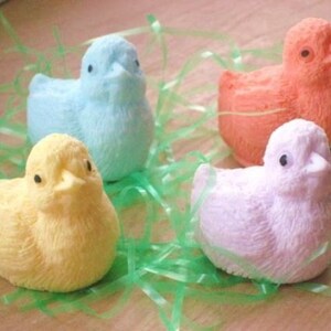 K&ouml;nnte beinhalten: Vier pastellfarbene Seifen in Form von K&uuml;ken, in den Farben Hellblau, Gelb, Rosa und Orange, liegen in einem Bett aus gr&uuml;nem, gras&auml;hnlichem Material.