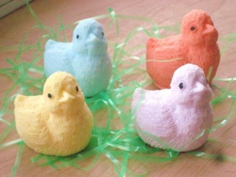 K&ouml;nnte beinhalten: Vier pastellfarbene Seifen in Form von K&uuml;ken, in den Farben Hellblau, Gelb, Rosa und Orange, liegen in einem Bett aus gr&uuml;nem, gras&auml;hnlichem Material.