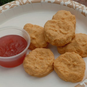 K&ouml;nnte beinhalten: Eine Gruppe von sechs goldbraunen, panierten H&auml;hnchen-Nuggets auf einem wei&szlig;en Pappteller mit beigem Blattmuster, begleitet von einem kleinen, durchsichtigen Plastikbeh&auml;lter, der mit einer durchscheinenden roten So&szlig;e gef&uuml;llt ist.