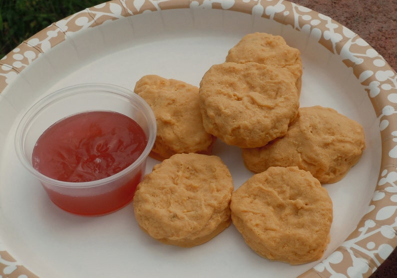 K&ouml;nnte beinhalten: Eine Gruppe von sechs goldbraunen, panierten H&auml;hnchen-Nuggets auf einem wei&szlig;en Pappteller mit beigem Blattmuster, begleitet von einem kleinen, durchsichtigen Plastikbeh&auml;lter, der mit einer durchscheinenden roten So&szlig;e gef&uuml;llt ist.