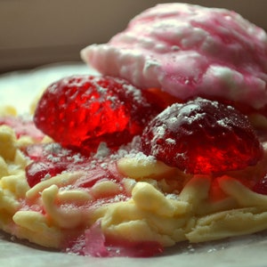 May include: A white cookie shaped like a pretzel with red and pink jelly candies on top. The cookie is covered in powdered sugar.
